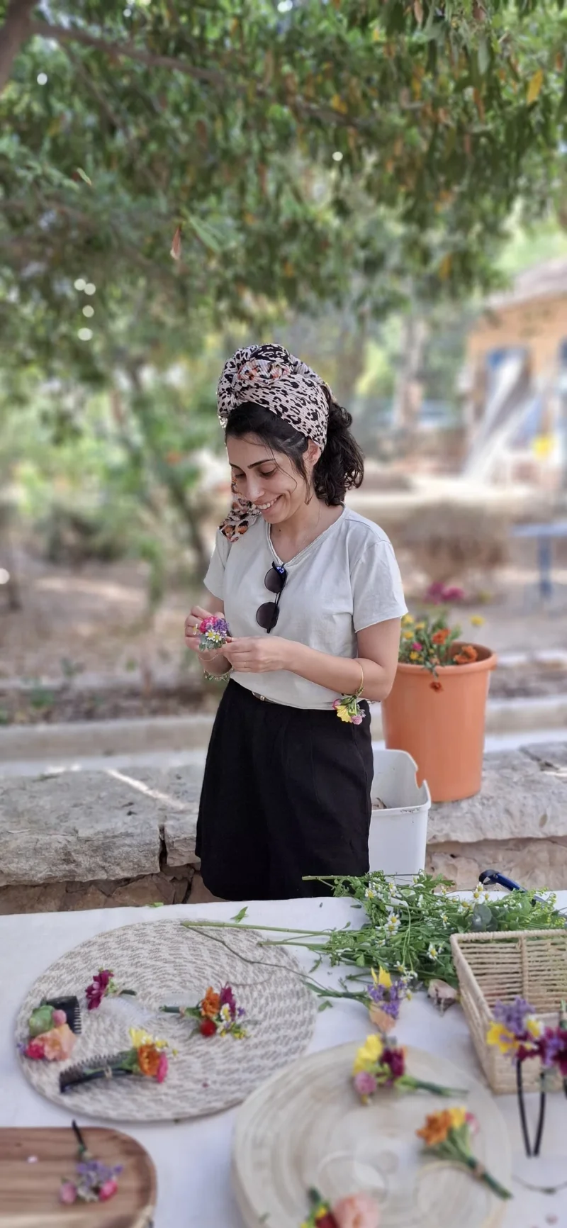 דנה מחייכת בכיסוי ראש מנומר, בזמן שזירת פרחים בגן